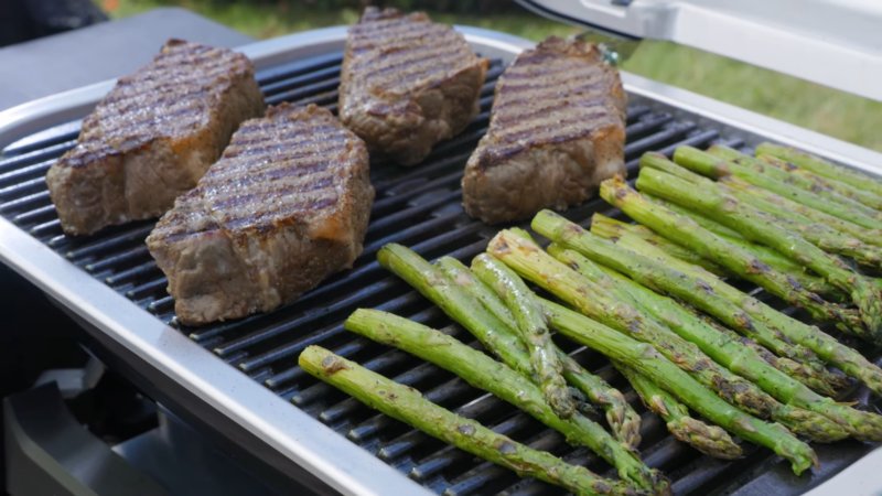 Dans cette image, nous voyons un barbecue électrique sur une terrasse en bois. Sur la grille, nous pouvons voir deux gros steaks juteux et des asperges qui grillent à la perfection. Les steaks sont bien marqués par la grille et les asperges sont légèrement dorées. On peut presque sentir l